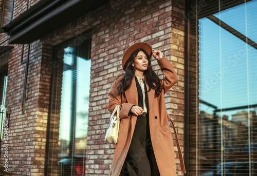 Asian business woman in long beige coat and hat walking down street in loft style, on background of brick wall, classic retro style