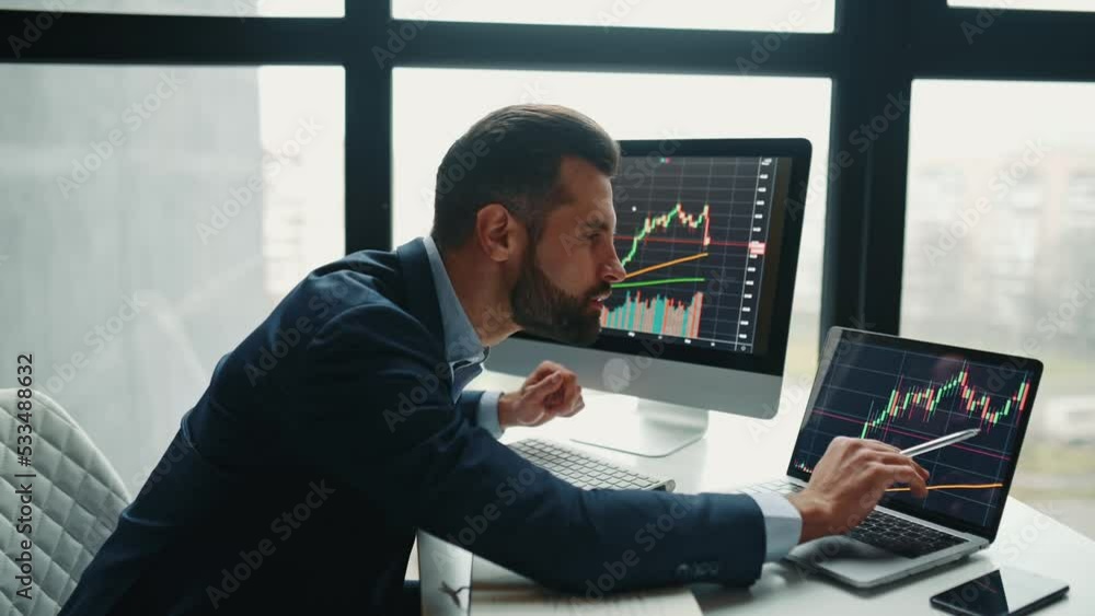 Focused smart successful caucasian male stock investor, broker, sits at his work desk, uses computer and laptop, concentrated analyzes risks and prospects, rise or fall of cryptocurrency coin