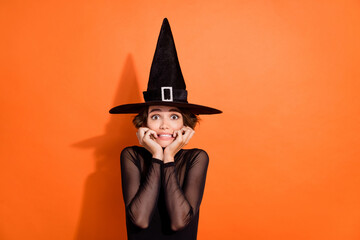 Photo portrait of gorgeous young girl scared bite nails bad news dressed trendy black halloween witch garment isolated on orange background