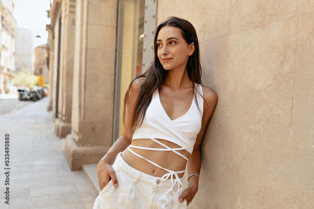 Attractive sling woman with long dark hair wearing summer light clothes looking aside near beige wall in summer sunny evening and smiling Attractive sling woman with long dark hair wearing summer light clothes looking aside near beige wall in summer sunny evening and smiling