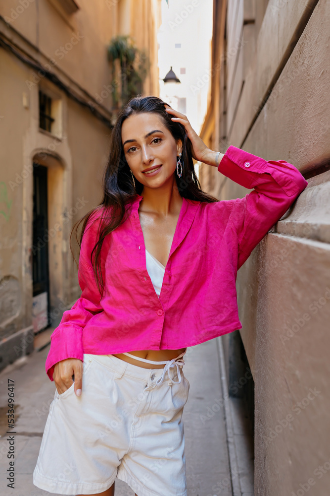 Smiling thin beautiful girl with loose long dark hair and wonderful smile wearing bright pink shirt posing on old european street