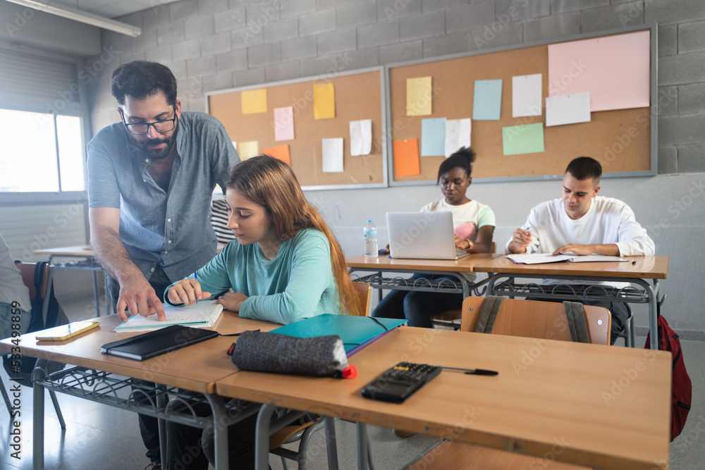 Young Professor helping female student in High School classroom. Friendly Teacher with beard explaining exercicse to blond girl