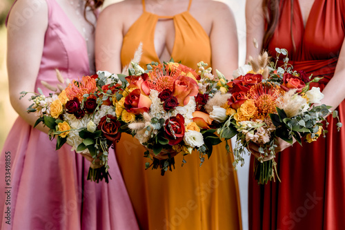 Bridesmaids in autumn colors holding colorful bouquets filled with orange roses and yellow anemones, close up crop no faces