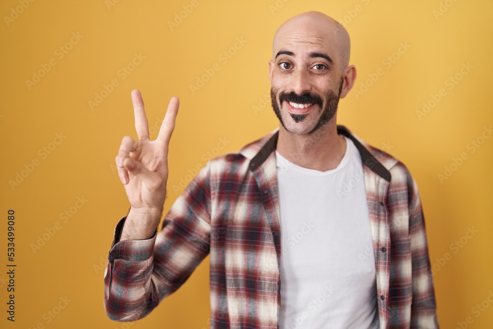 Naklejka premium Hispanic man with beard standing over yellow background showing and pointing up with fingers number two while smiling confident and happy.