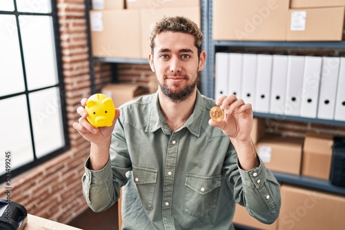 Hispanic man with beard working at small business ecommerce holding piggy bank and bitcoin relaxed with serious expression on face. simple and natural looking at the camera.