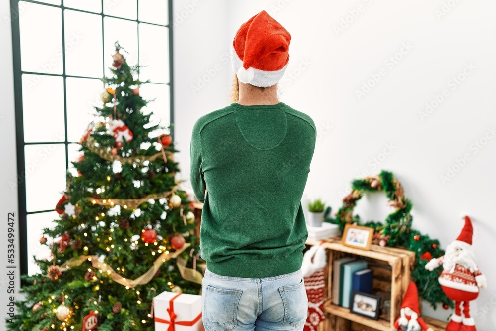 Redhead man with long beard wearing christmas hat by christmas tree standing backwards looking away with crossed arms Redhead man with long beard wearing christmas hat by christmas tree standing backwards looking away with crossed arms