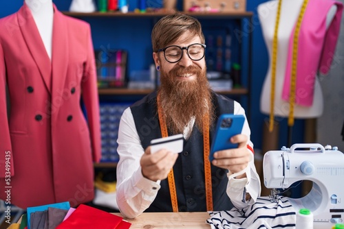 Young redhead man tailor using smartphone and credit card at clothing factory