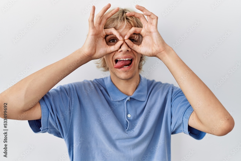 Young modern man standing over isolated background doing ok gesture like binoculars sticking tongue out, eyes looking through fingers. crazy expression. Young modern man standing over isolated background doing ok gesture like binoculars sticking tongue out, eyes looking through fingers. crazy expression.