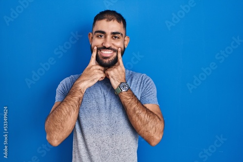 Middle east man with beard standing over blue background smiling with open mouth, fingers pointing and forcing cheerful smile