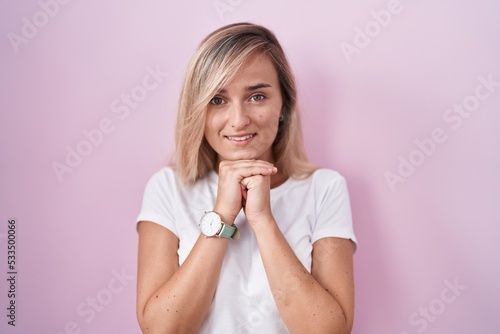 Young blonde woman standing over pink background laughing nervous and excited with hands on chin looking to the side