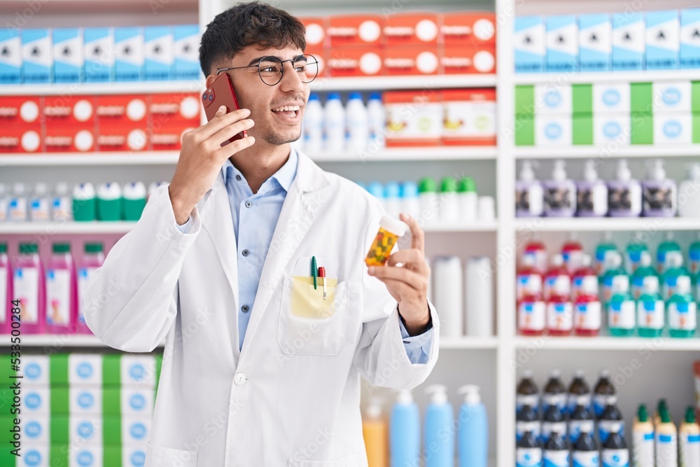 Young hispanic man pharmacist holding pills bottle talking on smartphone at pharmacy