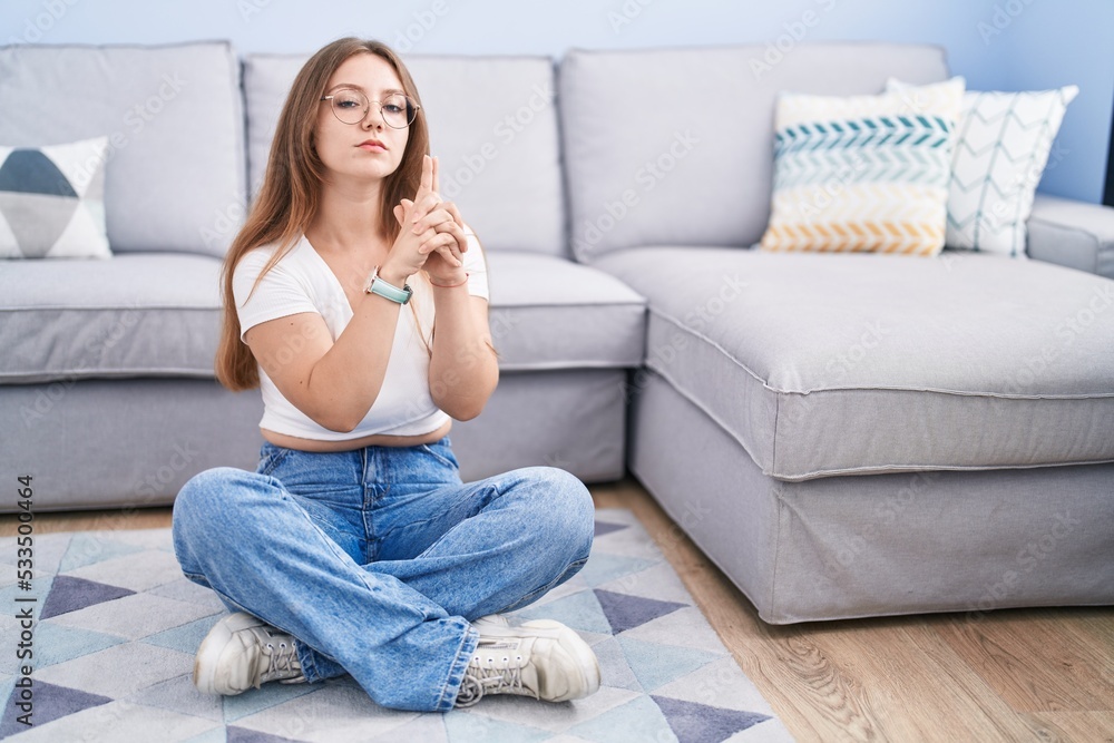 Young caucasian woman sitting on the floor at the living room holding symbolic gun with hand gesture, playing killing shooting weapons, angry face Young caucasian woman sitting on the floor at the living room holding symbolic gun with hand gesture, playing killing shooting weapons, angry face