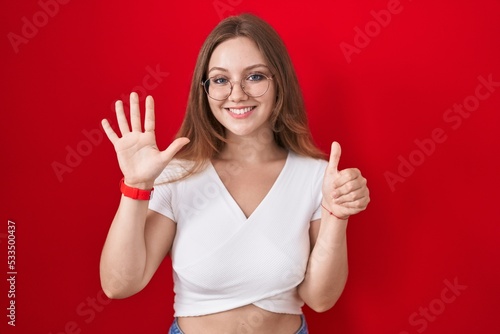 Young caucasian woman standing over red background showing and pointing up with fingers number six while smiling confident and happy.