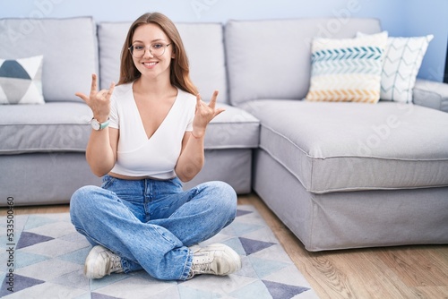 Young caucasian woman sitting on the floor at the living room shouting with crazy expression doing rock symbol with hands up. music star. heavy music concept.