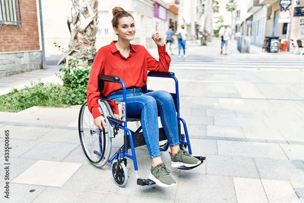 Young brunette woman sitting on wheelchair outdoors surprised with an idea or question pointing finger with happy face, number one Young brunette woman sitting on wheelchair outdoors surprised with an idea or question pointing finger with happy face, number one