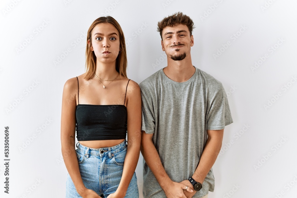 Naklejka premium Young beautiful couple standing together over isolated background puffing cheeks with funny face. mouth inflated with air, crazy expression.