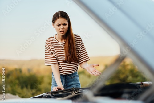 A sad woman has opened the hood of a broken down car and is looking for the cause of the breakdown on a road trip alone
