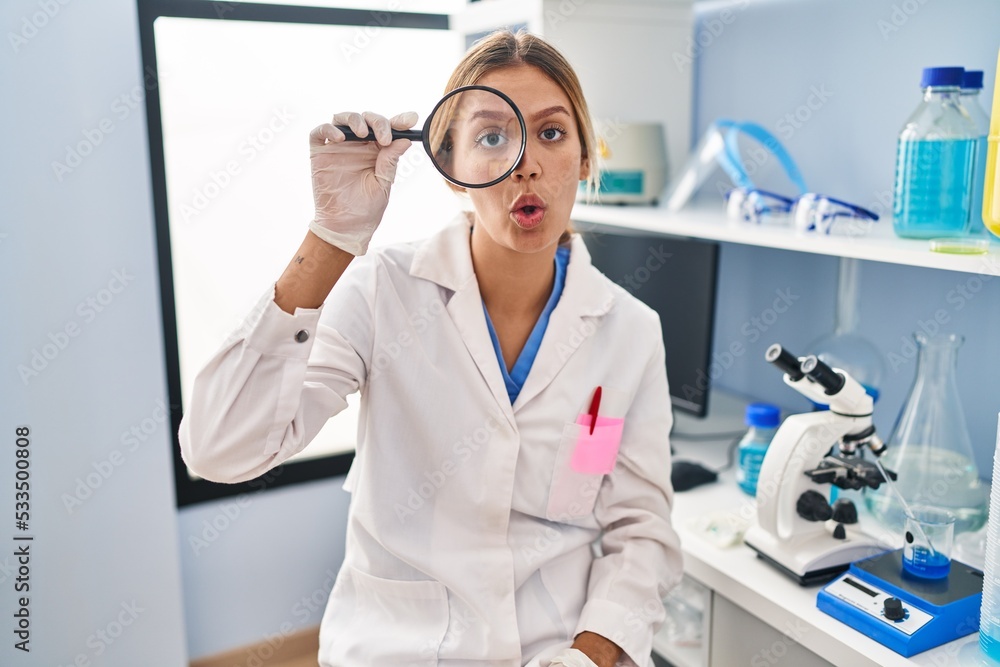 Young blonde woman working at scientist laboratory using magnifying glass scared and amazed with open mouth for surprise, disbelief face