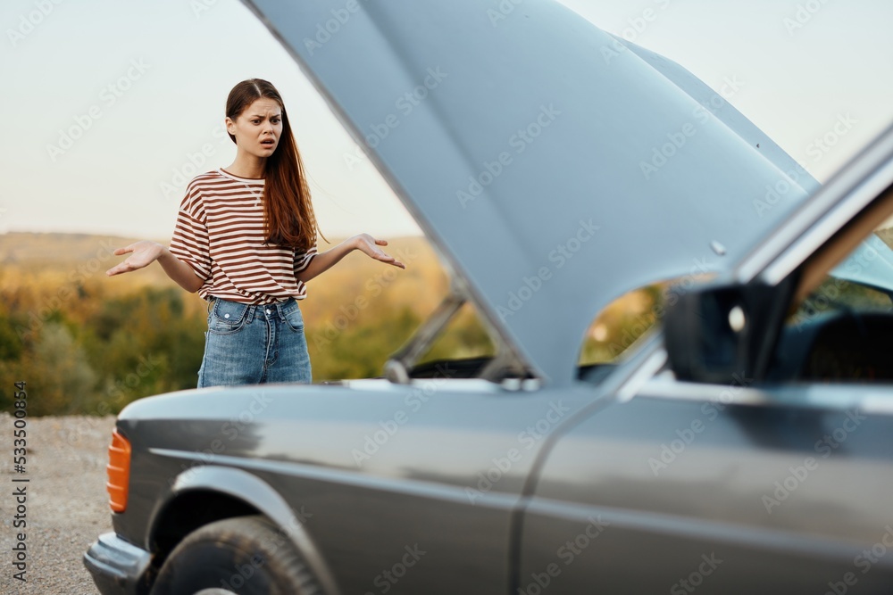 A sad woman has opened the hood of a broken down car and is looking for the cause of the breakdown on a road trip alone with her hands spread apart in incomprehension