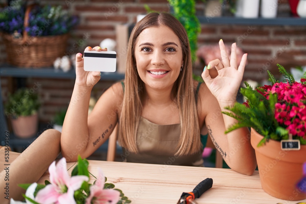 Young blonde woman working at florist shop holding credit card doing ok sign with fingers, smiling friendly gesturing excellent symbol