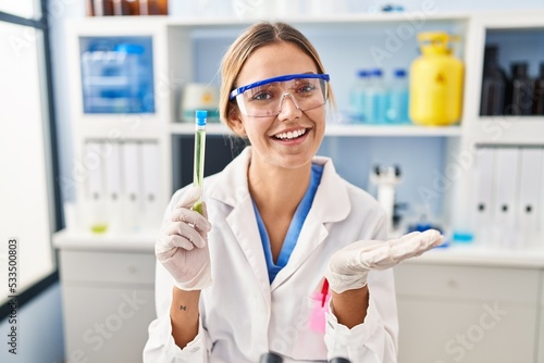 Young blonde woman working at scientist laboratory holding sample celebrating achievement with happy smile and winner expression with raised hand