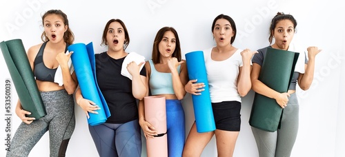 Group of women holding yoga mat standing over isolated background surprised pointing with hand finger to the side, open mouth amazed expression.