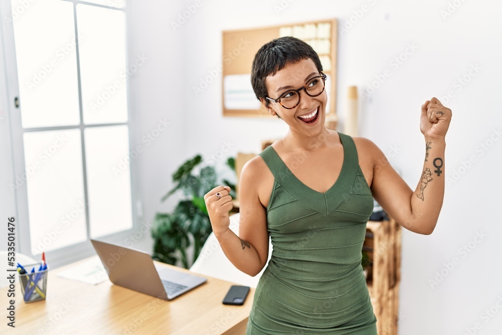 Young hispanic woman with short hair working at the office very happy and excited doing winner gesture with arms raised, smiling and screaming for success. celebration concept.