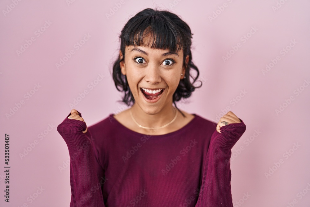 Young beautiful woman standing over pink background celebrating surprised and amazed for success with arms raised and open eyes. winner concept. Young beautiful woman standing over pink background celebrating surprised and amazed for success with arms raised and open eyes. winner concept.