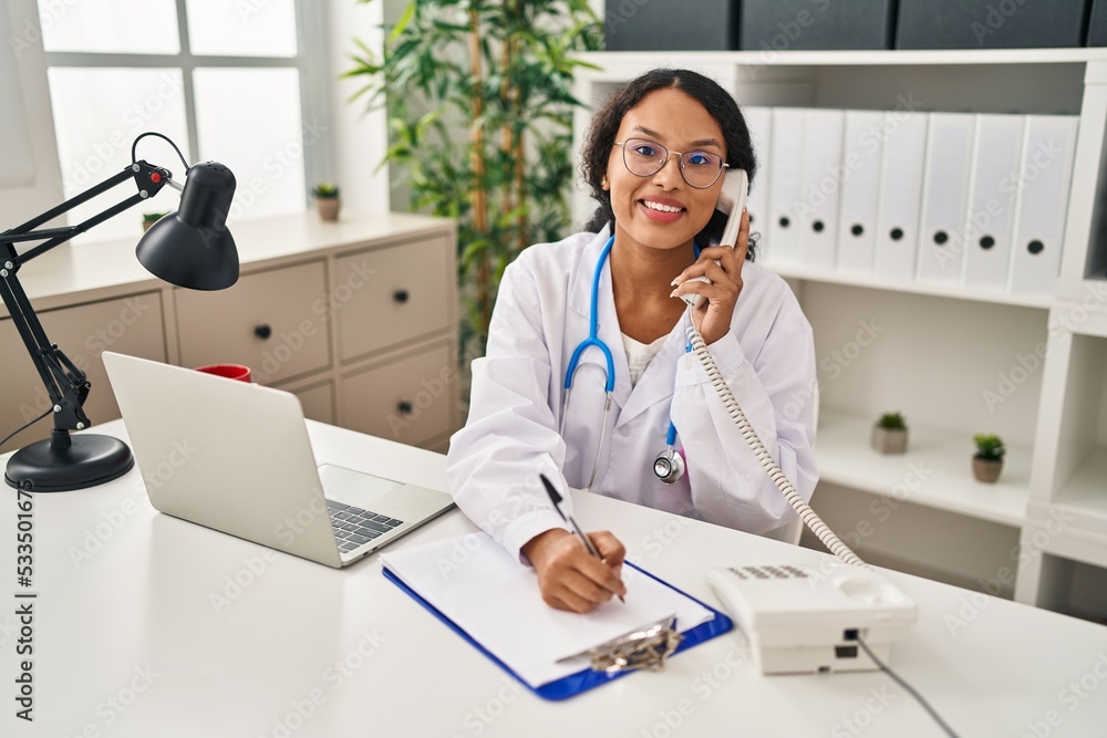 Young latin woman wearing doctor uniform talking on the telephone write on clipboard at clinic Young latin woman wearing doctor uniform talking on the telephone write on clipboard at clinic