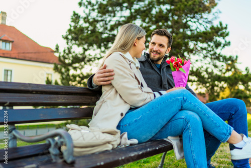 A young couple in love is sitting embraced on a park bench, they are talking and laughing, enjoying a beautiful sunny day.