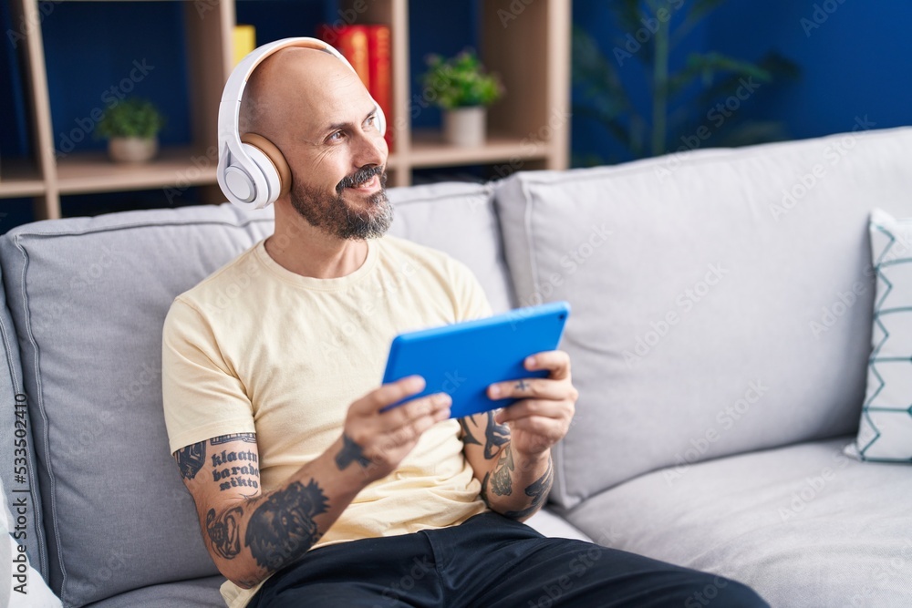Young bald man watching video on touchpad sitting on sofa at home