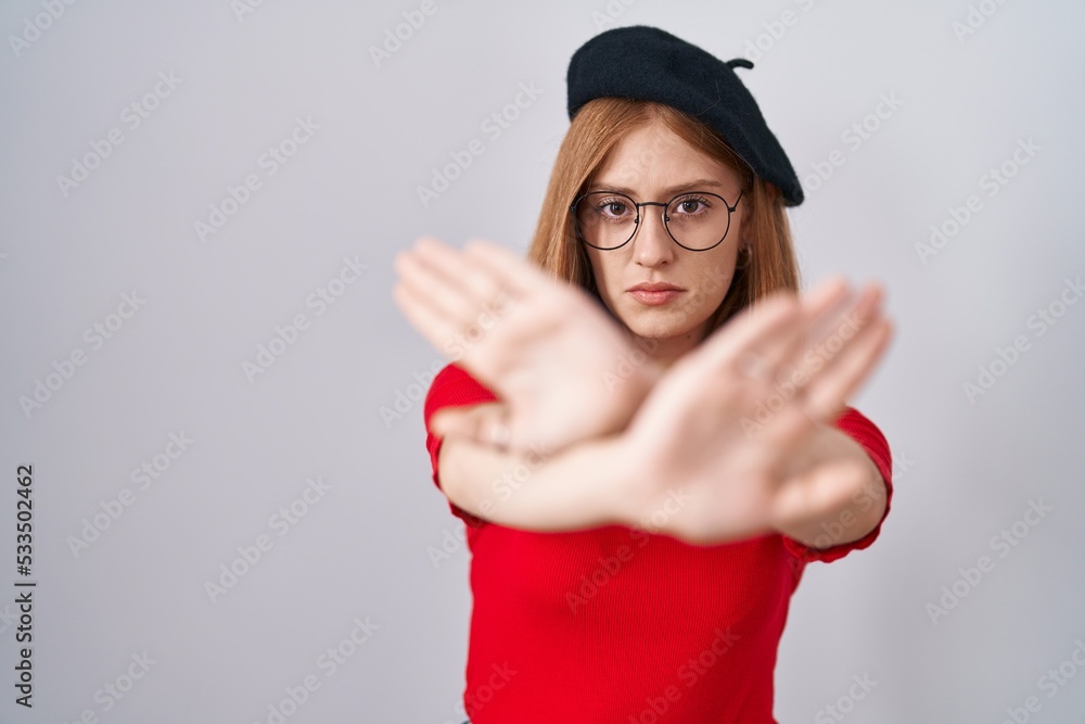 Young redhead woman standing wearing glasses and beret rejection expression crossing arms and palms doing negative sign, angry face