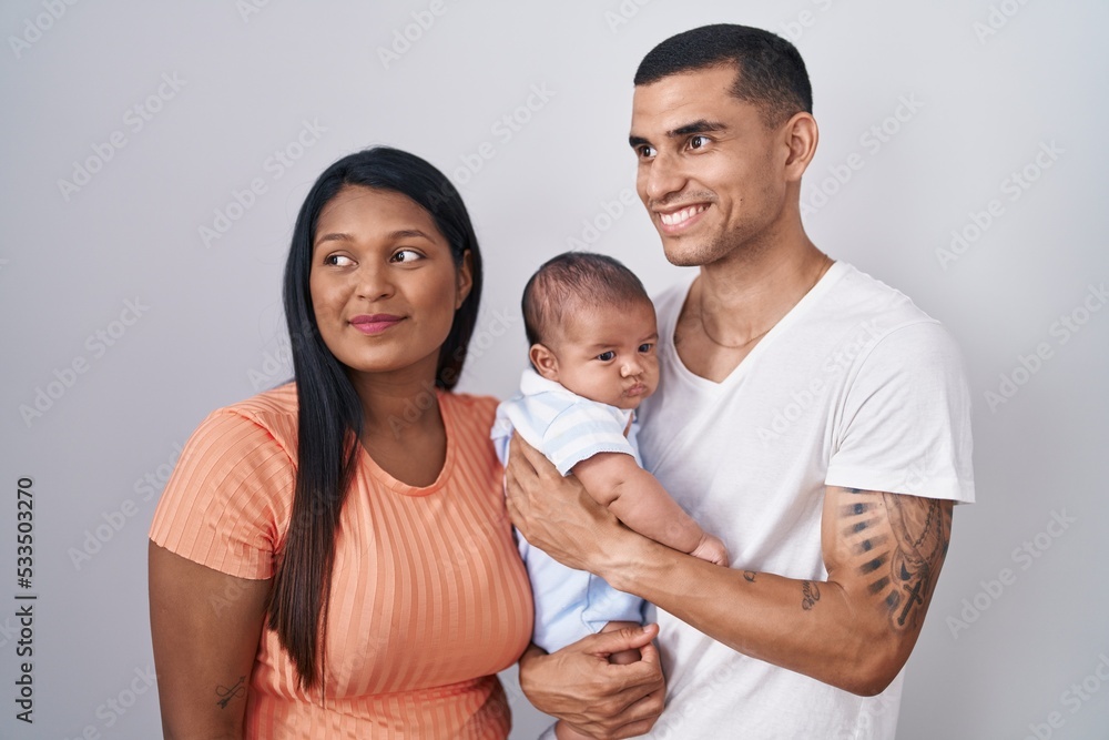 Young hispanic couple with baby standing together over isolated background looking away to side with smile on face, natural expression. laughing confident.
