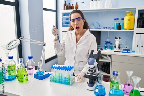 Young hispanic woman working at scientist laboratory scared and amazed with open mouth for surprise  disbelief face