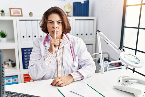 Middle age hispanic woman wearing doctor uniform and stethoscope at the clinic asking to be quiet with finger on lips. silence and secret concept.