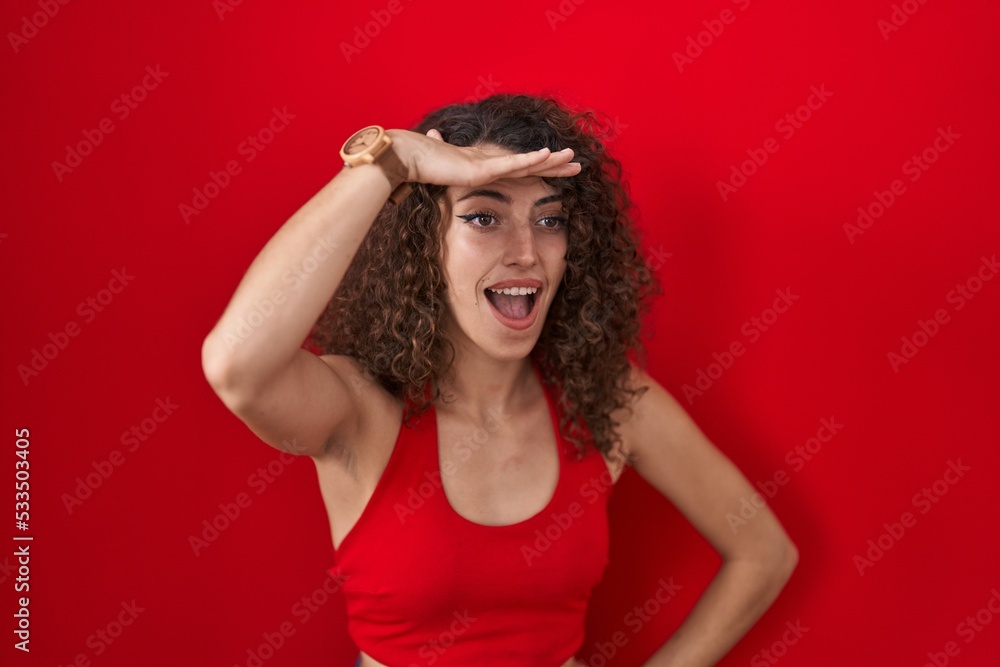 Hispanic woman with curly hair standing over red background very happy and smiling looking far away with hand over head. searching concept. Hispanic woman with curly hair standing over red background very happy and smiling looking far away with hand over head. searching concept.