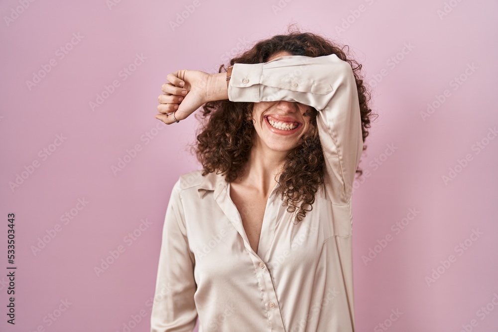 Naklejka premium Hispanic woman with curly hair standing over pink background covering eyes with arm smiling cheerful and funny. blind concept.
