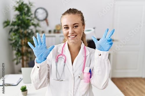 Young caucasian woman wearing doctor uniform and stethoscope at the clinic showing and pointing up with fingers number eight while smiling confident and happy.