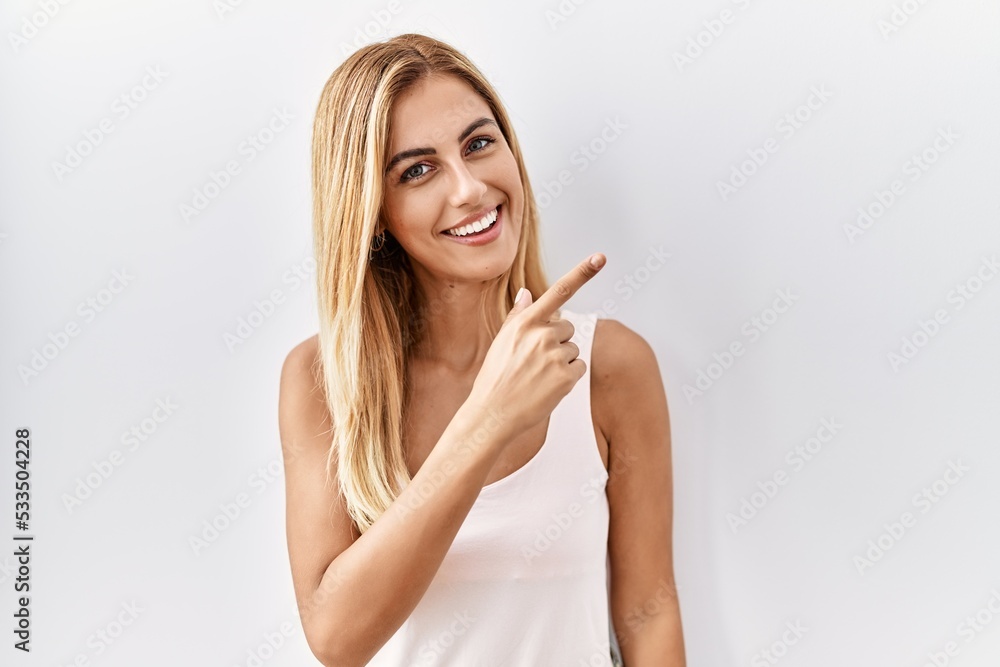 Blonde beautiful young woman standing over white isolated background cheerful with a smile of face pointing with hand and finger up to the side with happy and natural expression on face Blonde beautiful young woman standing over white isolated background cheerful with a smile of face pointing with hand and finger up to the side with happy and natural expression on face