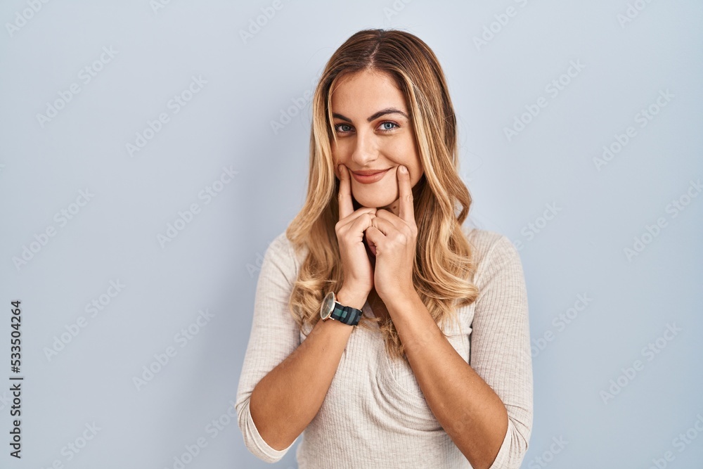 Young blonde woman standing over isolated background smiling with open mouth, fingers pointing and forcing cheerful smile Young blonde woman standing over isolated background smiling with open mouth, fingers pointing and forcing cheerful smile