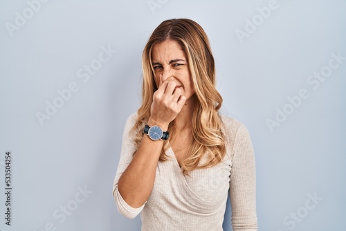 Young blonde woman standing over isolated background smelling something stinky and disgusting, intolerable smell, holding breath with fingers on nose. bad smell