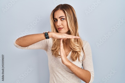 Young blonde woman standing over isolated background doing time out gesture with hands, frustrated and serious face