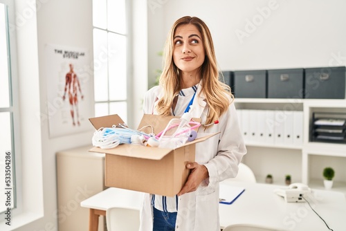 Young blonde doctor woman holding box with medical safety items smiling looking to the side and staring away thinking.