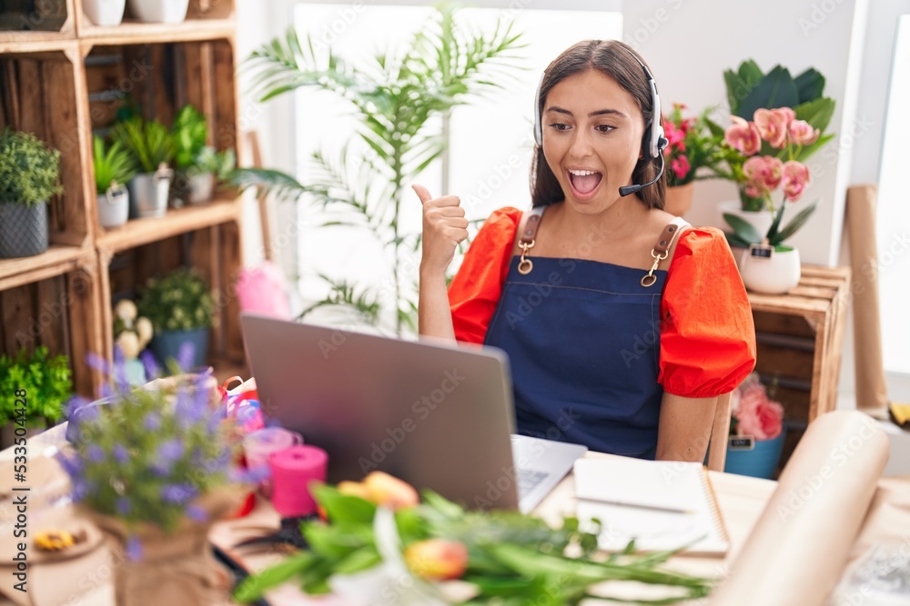 Young hispanic woman working at florist shop doing video call pointing thumb up to the side smiling happy with open mouth
