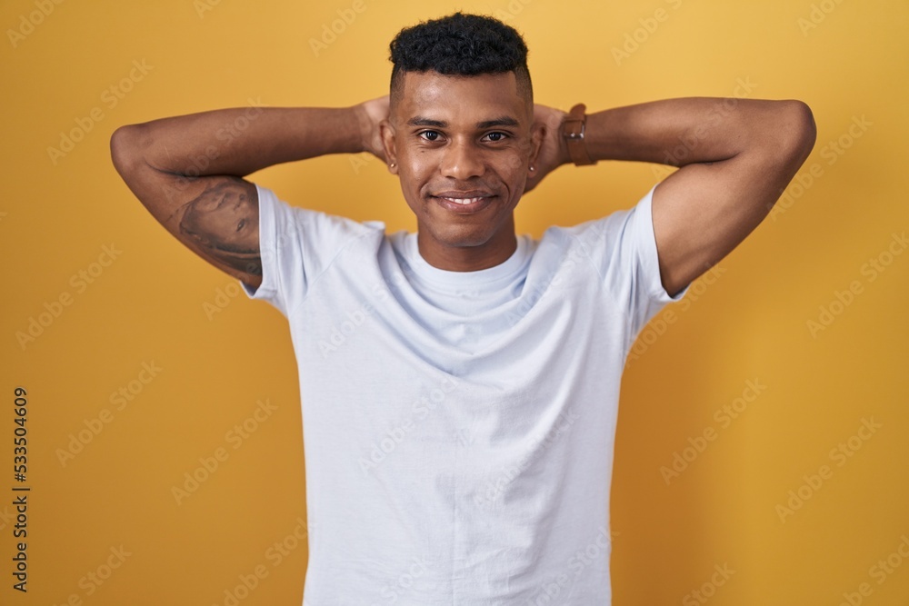 Young hispanic man standing over yellow background relaxing and stretching, arms and hands behind head and neck smiling happy Young hispanic man standing over yellow background relaxing and stretching, arms and hands behind head and neck smiling happy