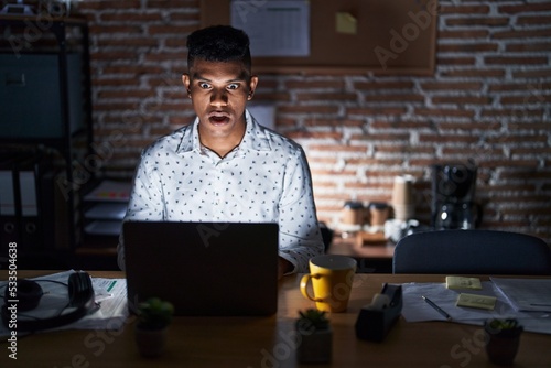 Young hispanic man working at the office at night afraid and shocked with surprise expression, fear and excited face.