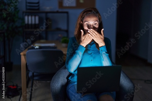 Brunette woman working at the office at night shocked covering mouth with hands for mistake. secret concept.
