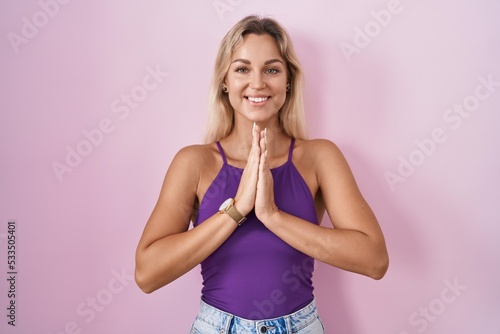 Young blonde woman standing over pink background praying with hands together asking for forgiveness smiling confident.