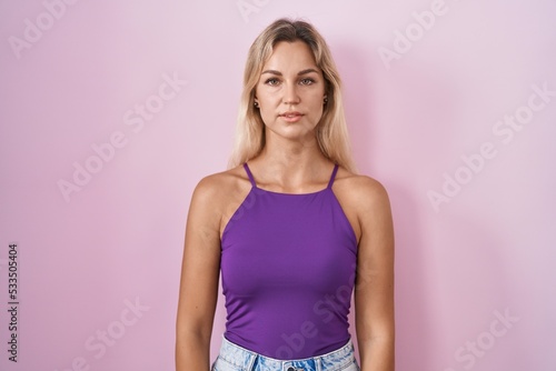 Young blonde woman standing over pink background relaxed with serious expression on face. simple and natural looking at the camera.