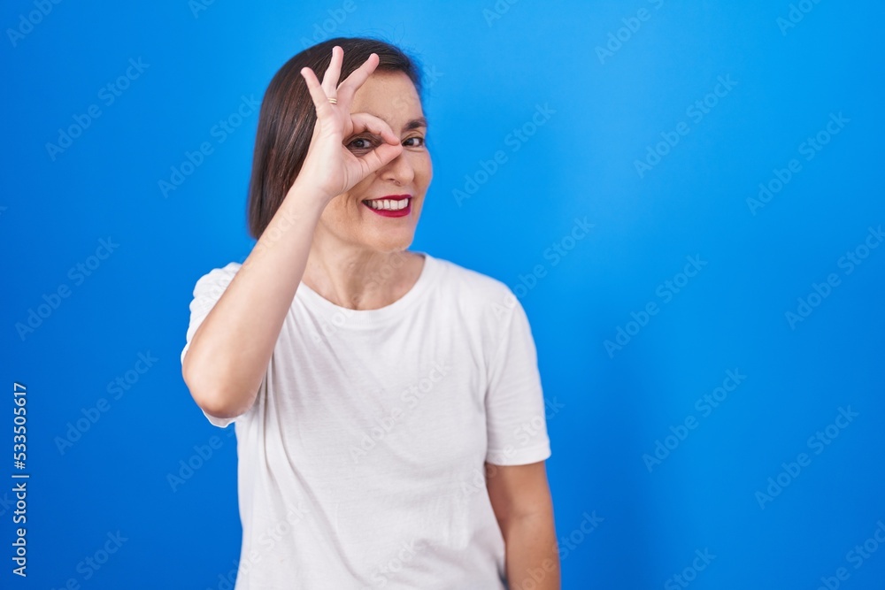 Middle age hispanic woman standing over blue background doing ok gesture with hand smiling, eye looking through fingers with happy face. Middle age hispanic woman standing over blue background doing ok gesture with hand smiling, eye looking through fingers with happy face.